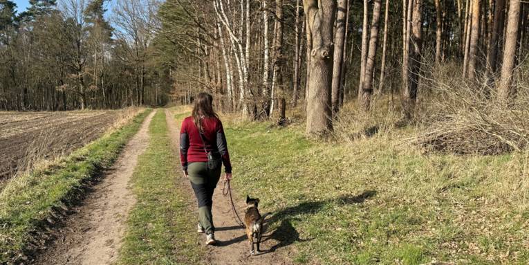 Eine Frau führt einen Hund an der Leine auf einem Weg zwischen Feld und Wald.