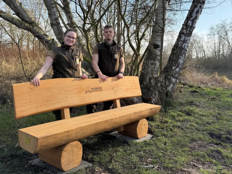 Eine Naturpark-Rangerin und eine Naturpark-Ranger stehen hinter einer Holzbank.