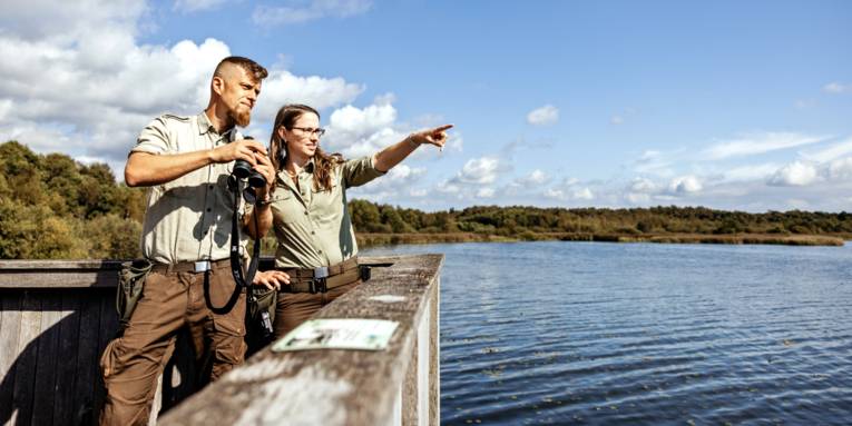 Eine Rangerin und ein Ranger stehen auf einer Aussichtsplattform. Er h&auml;lt ein Fernglas in den H&auml;nden, sie zeigt mit dem Finger ein eine Richtung.