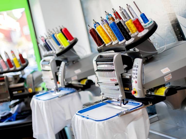 Stoff ist in einen Rahmen gespannt, eine Maschine bestickt den gespannten Stoff innerhalb des Rahmens. Auf der Maschine sind verschiedenfarbige Garnrollen.