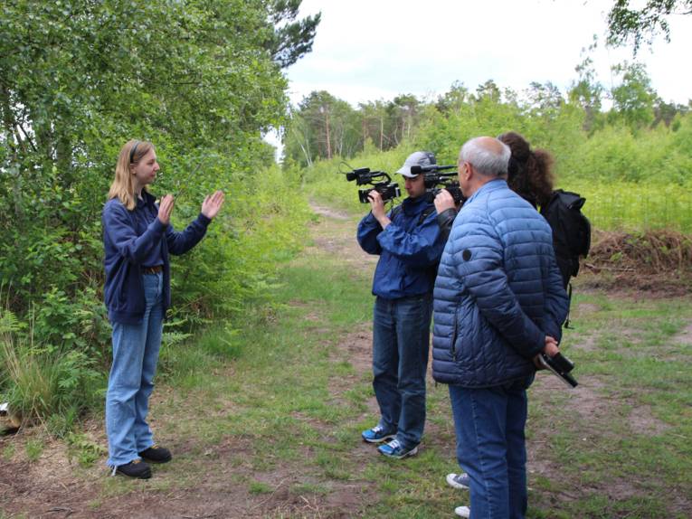 Eine Frau steht in einer Moorlandschaft. Zwei Männer halten jeweils eine Kamera auf sie, ein weiterer Mann steht daneben.