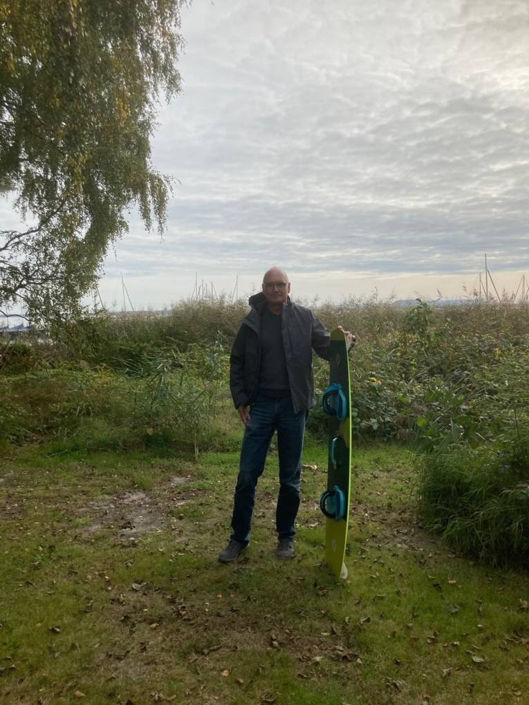 Ein Mann steht neben einem hochkant stehenden Kiteboard vor einem See
