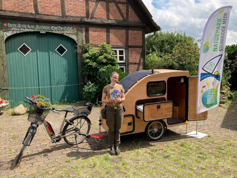 Eine Frau trägt Outdoor-Kleidung, hält Infomaterial in ihren Händen und steht vor einem besonderen Gespann aus E-Bike und Fahrradanhänger, einem kleinem Wohnmobil aus Holz.