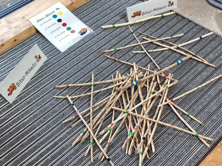 Holzstöckchen unterschiedlicher Beschaffenheit sind jeweils farblich markiert und liegen in einem wilden Haufen aufeinander. Papieraufsteller tragen die Aufschrift "Biber-Mikado", eine Tafel liegt auf dem Boden zwischen den Papieraufstellern und gibt an, welche Farbe in diesem rustikalen Mikadospiel welchen Punktewert hat.