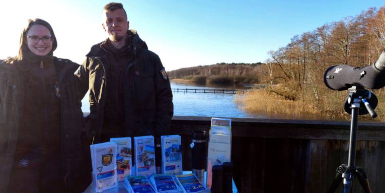 Zwei Personen tragen Kleidung für draußen und stehen auf einer Aussichtsplattform. Auf einer Kiste vor ihnen ist Informationsmaterial. Im Hintergrund ist das Steinhuder Meer.