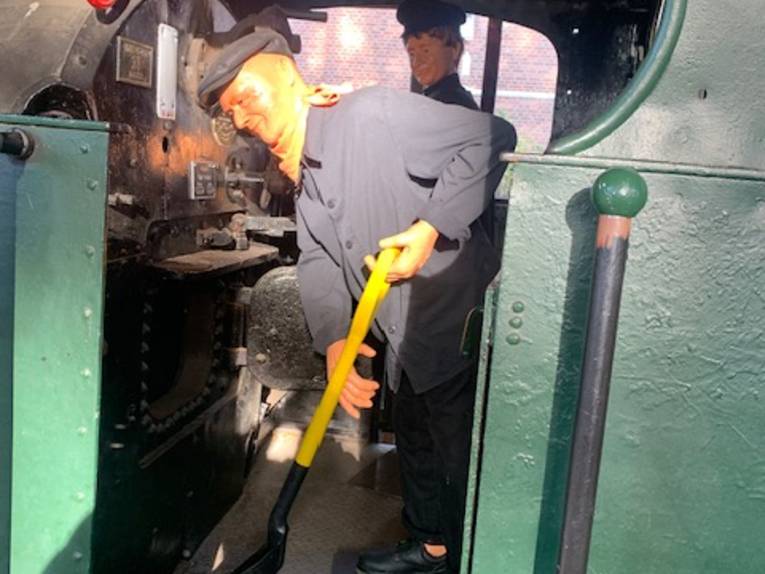 Blick auf den Führerstand einer historischen Eisenbahn, eine Person schaufelt Kohlen.