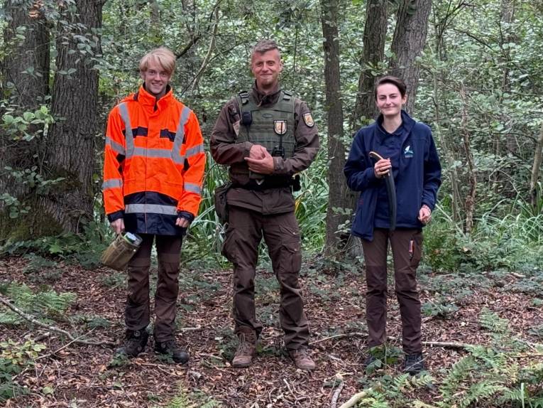 Drei Personen stehen in einem Wald und tragen Kleidung, Arbeitskleidung.