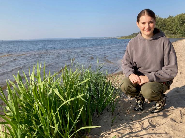 Eine Person hockt an einem Strand neben Pflanzen.
