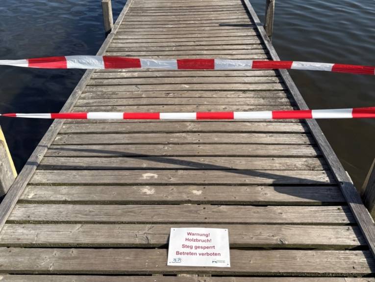 Rot-weißes Absperrband versperrt den Zugang zu einem Steg. Auf dem Steg ist ein Schild: "Warnung! Holzbruch! Steg gesperrt | Betreten verboten"