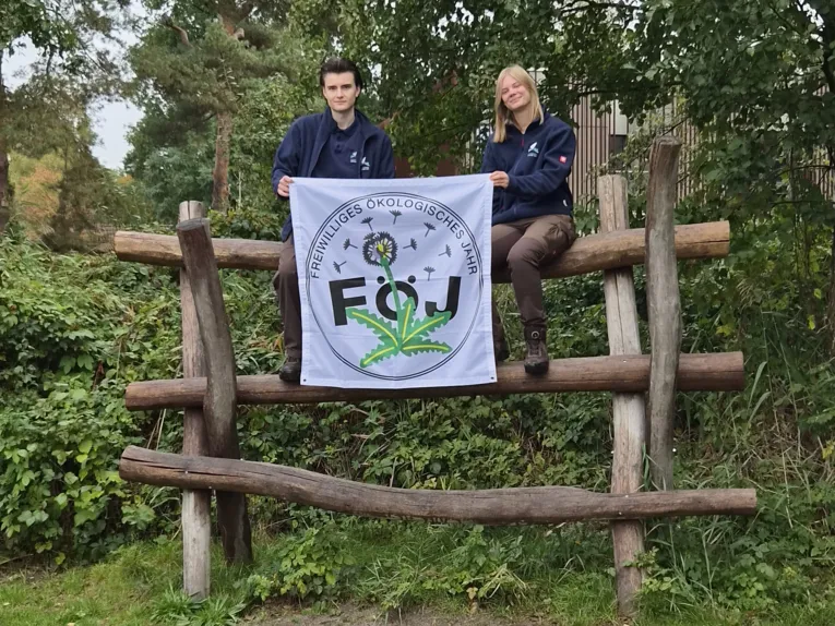 Zwei Personen sitzen auf einem Baumstamm und halten eine quadratische Fahne, auf der steht "Freiwilliges Ökologisches Jahr | FÖJ