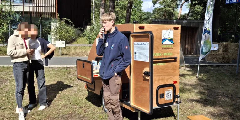 Ein Mann tr&auml;gt Outdoor-Kleidung, steht vor einem besonderen Fahrradanh&auml;nger aus Holz mit Mikrofontechnik und Infomaterial, er spricht in ein Mikrofon.