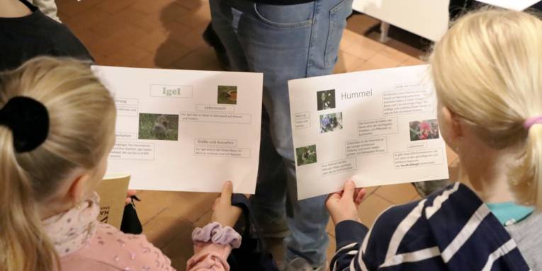 Zwei Mädchen halten jeweils ein Blatt Papier mit Informationen: Links über den Igel, rechts über die Hummel.