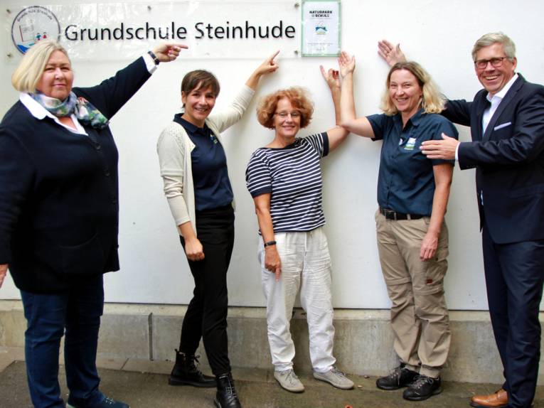 Vier Frauen und ein Mann zeigen auf ein teilweise transparentes Schild an einem Schulgebäude. Auf dem Schild steht unter anderem "Grundschule Steinhude" und "Naturparkschule".