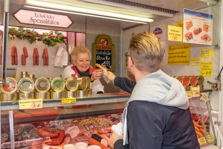 Eine Verkäuferin reicht einem Kunden eine Bockwurst.