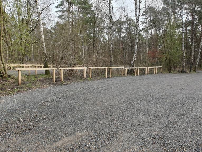 Blick auf einen geschotterten Parkplatz, Holzgeländer grenzen die Parkflächen von nahen Baumbestand ab.