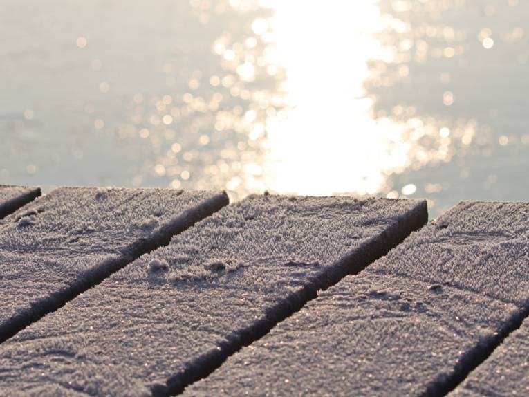 Blick über einen von Eis überzogenen Steg. Hinter und unter den vereisten Holzbrettern schimmert eine von der Sonne beschienene Wasserfläche.