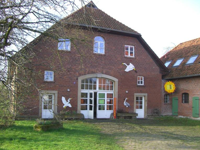 Älteres Backsteingebäude mit angrenzender Stallung. An der Fassade sind Bilder von Reihern und einer Sonne befestigt