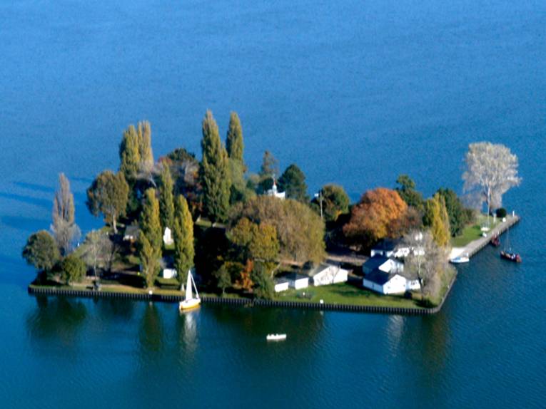 Luftaufnahme: Nahezu quadratische, flache Insel mit Bäumen und Gebäuden. Am Ufer ankern zwei Segelbote.
