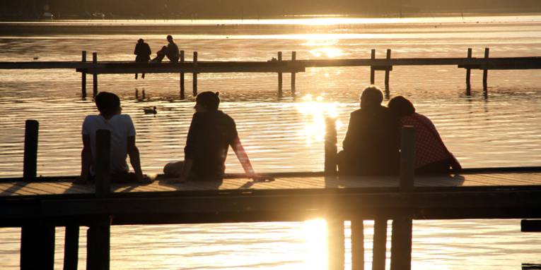 Zwei Steege ragen ins Steinhuder Meer, darauf sitzen sechs Menschen während die Sonne untergeht.