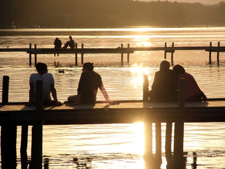 Zwei Steege ragen ins Steinhuder Meer, darauf sitzen sechs Menschen während die Sonne untergeht.