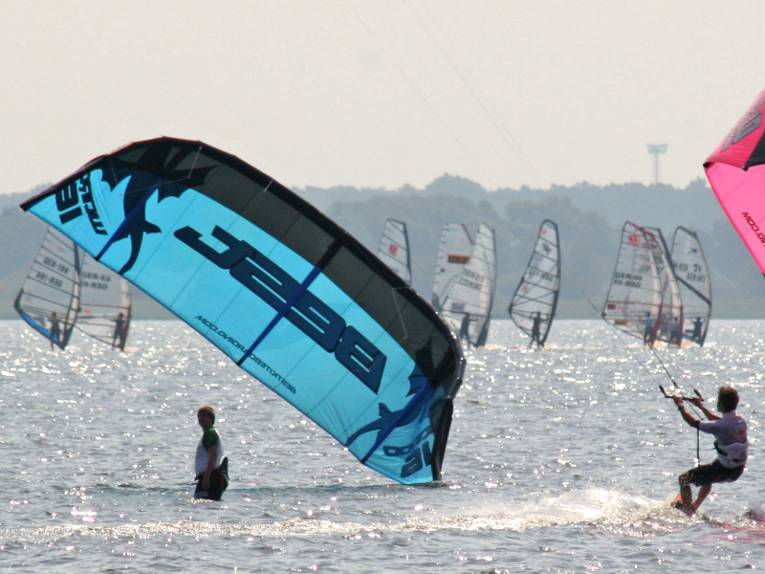 Eine Person steht auf einem Surfbrett und lässt sich von einem Lenkdrachen über das Wasser ziehen. Links ist ein blauer Lenkdrachen eines weiteren Kitesurfers zu erkennen, im Hintergrund sind Wassersportler als Windsurfer unterwegs.