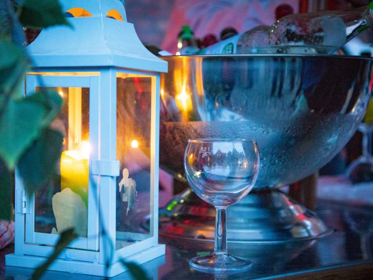 In einer Laterne leuchtet eine Kerze. Daneben liegt eine Flasche Mineralwasser auf Eis in einem Flaschenkühler, ein leeres Wasserglas steht bereit.