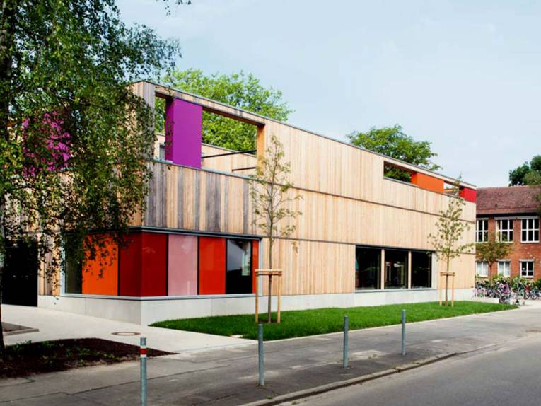 Die neugebaute Mensa der Grundschule Entenfang ist ein holzverkleideter Kubus mit barrierefreiem Zugang und bunten Tür- und Fensterelementen in orange, fliederfarben und rot.