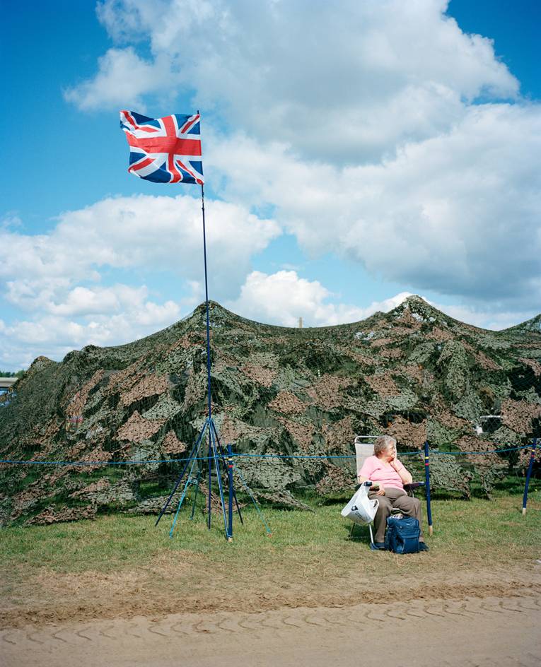 Eine Frau sitzt auf einem Stuhl vor einem großen militärischen Tarnnetz, an einem Mast weht die Flagge Großbritanniens.