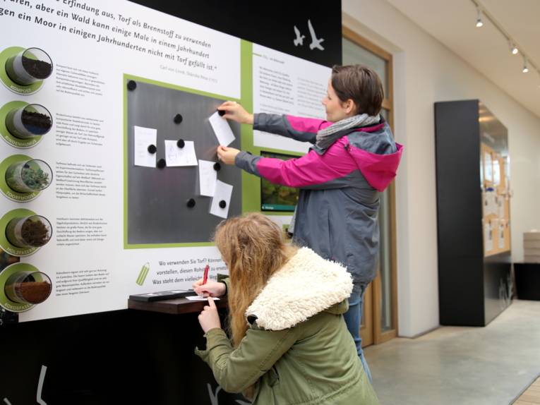 Zwei Frauen schreiben Kärtchen und hängen sie an eine Magnettafel. In Schaugläsern sind fünf Naturmaterialien zu sehen, zum Beispiel Torfmosse, Holzfasern und Rindenhumus.