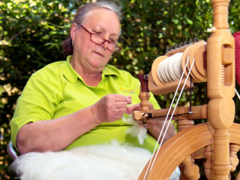 Eine Frau sitzt an einem Spinnrad und spinnt grobe Wolle zu einem gleichmäßigen Faden.