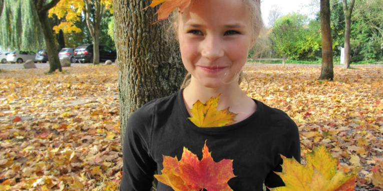 Ein Mädchen steht unter einem Baum. Herbstlaub liegt auf dem Fußboden, auch auf dem Kopf des Mädchens liegt ein Blatt und es hält Blätter in den Händen.