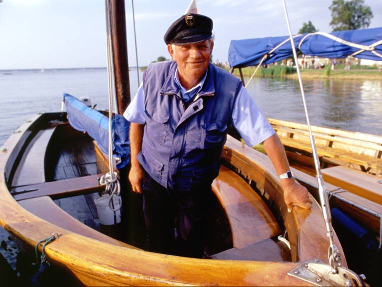 Ein Mann mit Schiffermütze und maritimer Kleidung steht in einem Boot des Typs "Auswanderer", einer offenen Segeljolle, welche als Ausflugsboot auf dem Steinhuder Meer unterwegs ist.