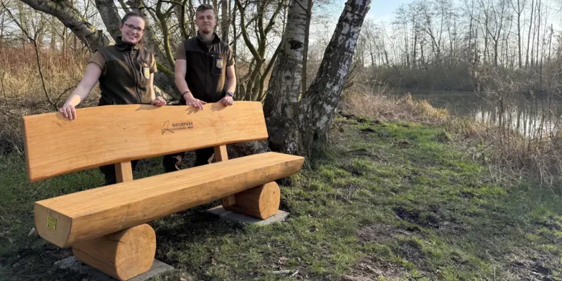 Eine Naturpark-Rangerin und eine Naturpark-Ranger stehen hinter einer Holzbank.