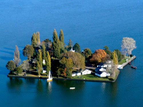 Luftaufnahme: Nahezu quadratische, flache Insel mit Bäumen und Gebäuden. Am Ufer ankern zwei Segelbote. Luftaufnahme: Nahezu quadratische, flache Insel mit Bäumen und Gebäuden. Am Ufer ankern zwei Segelbote.