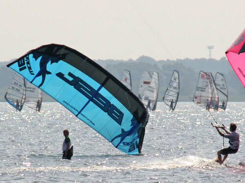 Eine Person steht auf einem Surfbrett und lässt sich von einem Lenkdrachen über das Wasser ziehen. Links ist ein blauer Lenkdrachen eines weiteren Kitesurfers zu erkennen, im Hintergrund sind Wassersportler als Windsurfer unterwegs. Eine Person steht auf einem Surfbrett und lässt sich von einem Lenkdrachen über das Wasser ziehen. Links ist ein blauer Lenkdrachen eines weiteren Kitesurfers zu erkennen, im Hintergrund sind Wassersportler als Windsurfer unterwegs.