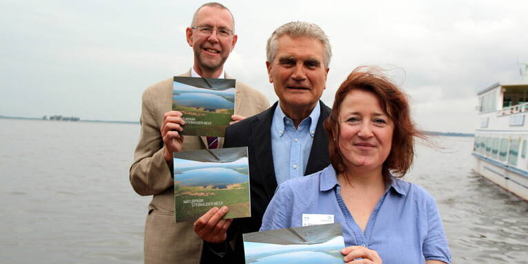 Prof. Dr. Axel Priebs (von links), Umweltdezernent der Region Hannover, Dr. Michael Arndt, Präsident des Verbandes deutscher Naturparke und Andrea Zörnig, Geschäftsführerin des Naturparks Steinhuder Meer stehen versetzt hintereinander am Ufer des Steinhuder Meeres. Jede Person hält eine Ausgabe des Buches "Naturpark Steinhuder Meer" in den Händen. Im Hintergrund sind die Festung Wilhelmstein und ein Ausflugsboot zu erkennen. Prof. Dr. Axel Priebs (von links), Umweltdezernent der Region Hannover, Dr. Michael Arndt, Präsident des Verbandes deutscher Naturparke und Andrea Zörnig, Geschäftsführerin des Naturparks Steinhuder Meer stehen versetzt hintereinander am Ufer des Steinhuder Meeres. Jede Person hält eine Ausgabe des Buches "Naturpark Steinhuder Meer" in den Händen. Im Hintergrund sind die Festung Wilhelmstein und ein Ausflugsboot zu erkennen.