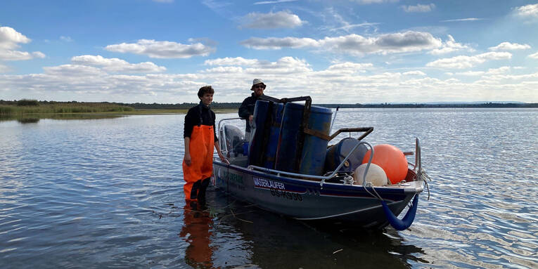 Ein Mann trägt eine orangefarbene Wathose und steht neben einem Boot. In dem Boot ist ein zweiter Mann und jede Menge Unrat einer Müllsammelaktion. Ein Mann trägt eine orangefarbene Wathose und steht neben einem Boot. In dem Boot ist ein zweiter Mann und jede Menge Unrat einer Müllsammelaktion.