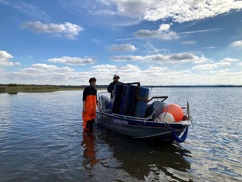 Ein Mann trägt eine orangefarbene Wathose und steht neben einem Boot. In dem Boot ist ein zweiter Mann und jede Menge Unrat einer Müllsammelaktion. Ein Mann trägt eine orangefarbene Wathose und steht neben einem Boot. In dem Boot ist ein zweiter Mann und jede Menge Unrat einer Müllsammelaktion.