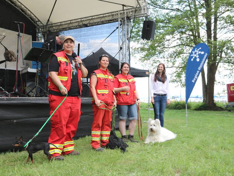 Vier Menschen und drei Hunde stehen vor einer Bühne. Vier Menschen und drei Hunde stehen vor einer Bühne.