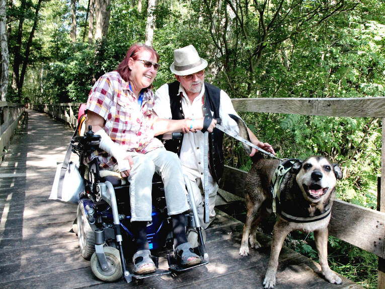 Eine Frau, ein Mann und ein Hund sind gemeinsam auf einem Holzsteg, die Frau fährt einen Rollstuhl und der Mann führt den Hund an der Leine. Für das Foto posieren sie beieinander.
Eine Frau, ein Mann und ein Hund sind gemeinsam auf einem Holzsteg, die Frau fährt einen Rollstuhl und der Mann führt den Hund an der Leine. Für das Foto posieren sie beieinander.